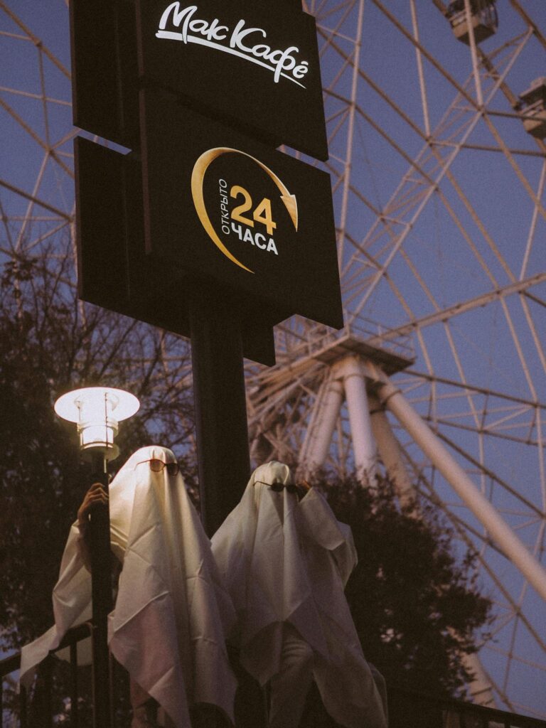 Two ghostly figures stand under a streetlight near a Ferris wheel on a Halloween evening.